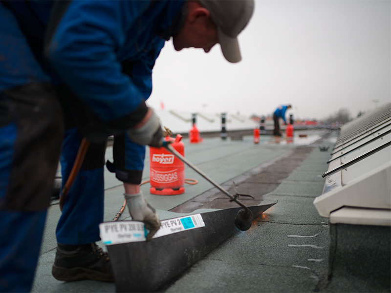 Dachdecker klebt schwarze Bitumen-Dachpappe auf Flachdach bei Dachabdichtung prüfen zur sicheren Abdichtung
